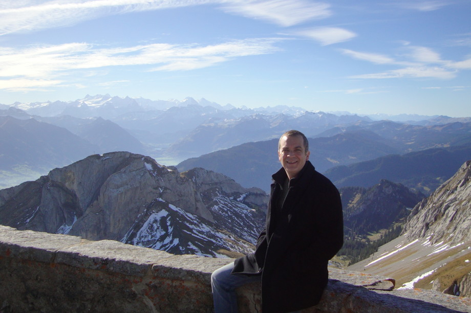 Me sitting on a waist-level wall on a sunny Mt Pilatus in Switzerland, with mountains in the background