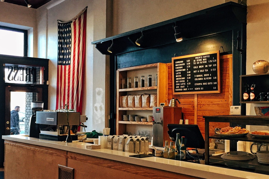 Cash register and counter of a coffee shop with various colored woods and an American flag hanging on the wall, and lots of pastries and bags of coffee beans