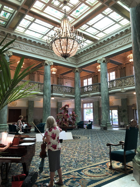 Musicians playing in a foyer with green marble columns, a chandelier, and skylight.