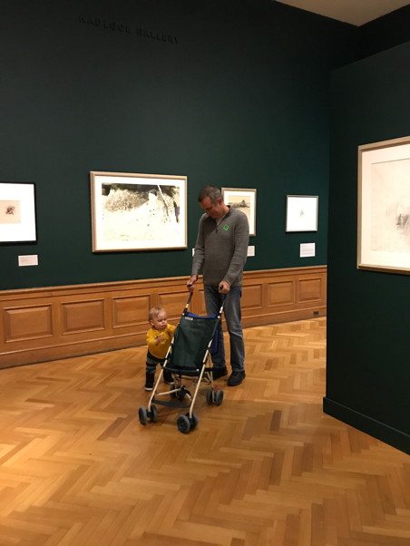 Child rocking an empty stroller that dad is pushing through a gallery with green walls and polished wooden floors