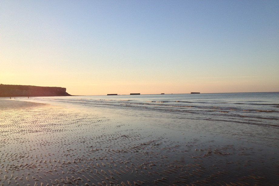 An empty beach at sunset, with a cliff in the distance and large dashes along the horizon that look like elongated train cars