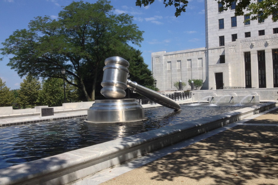An enormous stainless steel gavel strikes a stainless steel sound block in the middle of a pool on a brilliant summer day