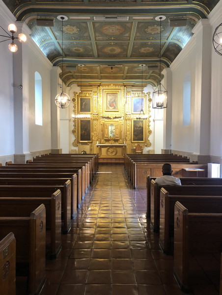 The interior of a modest yet beautiful church—spotless earthen tile floor, immaculate white walls, kaleidoscope-patterned ceiling, and enormous gold-framed paintings behind the altar