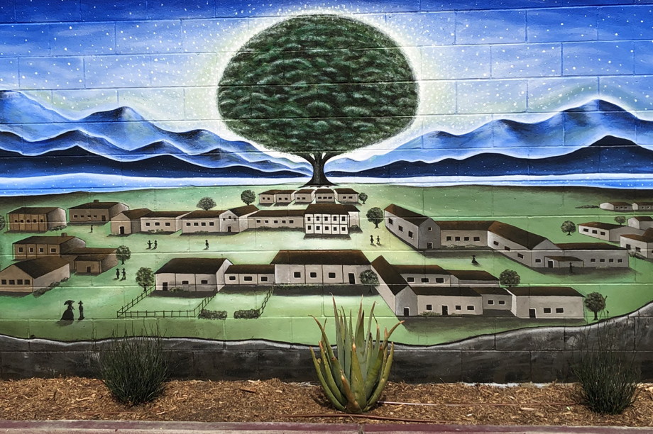 A mural of the pueblo de Los Angeles, with an illuminated tree, blue skies, and small buildings in a green valley