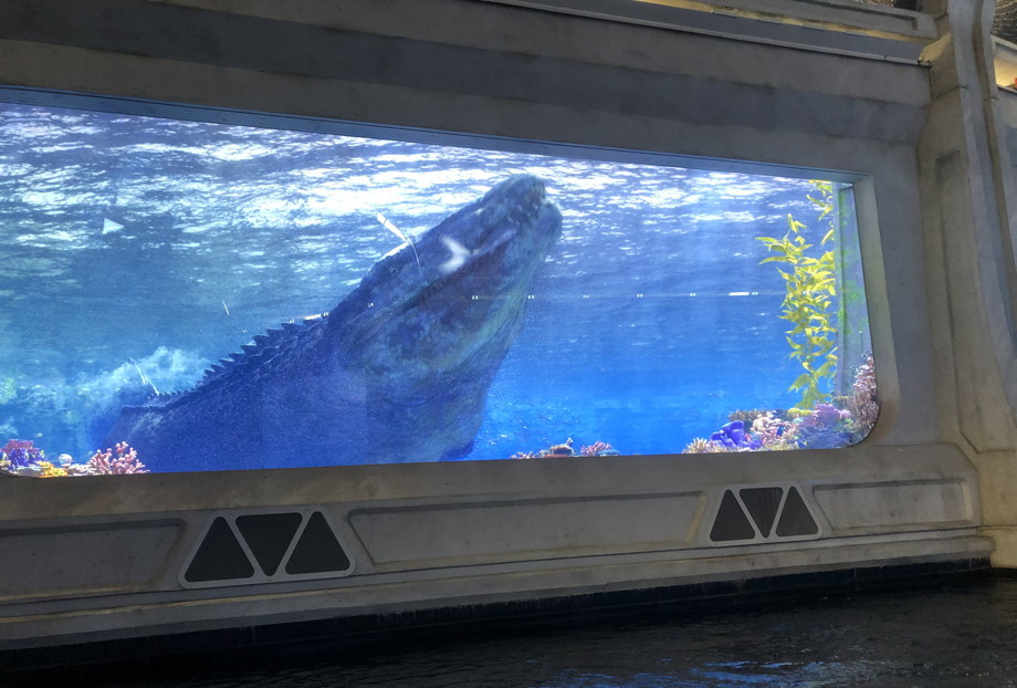 A window with blue water and plants, with a mosasaurus swimming around, it looks like a whale-sized crocodile