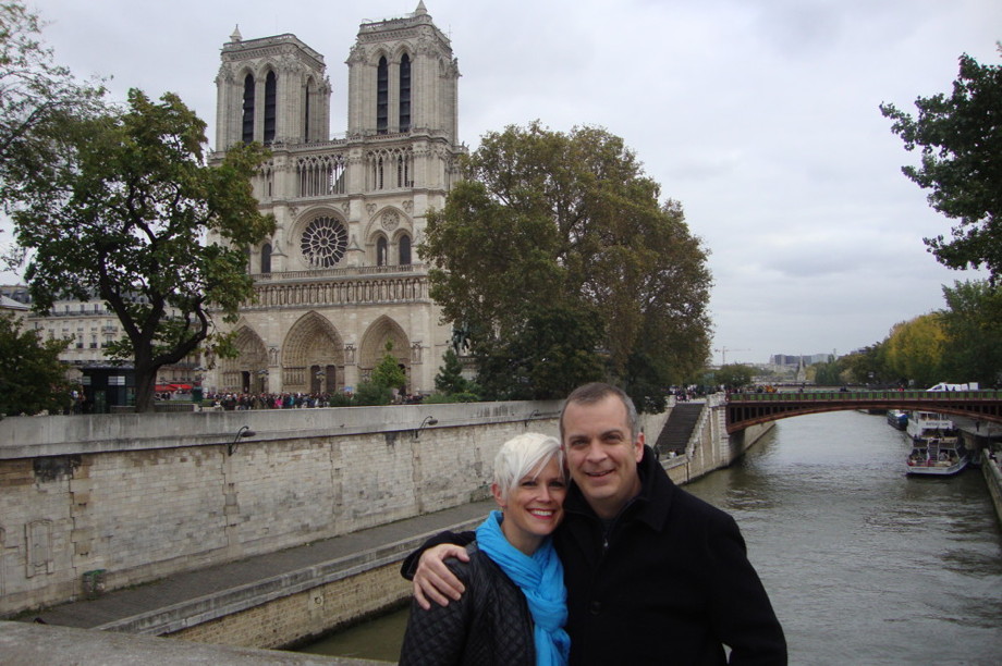 A couple standing in front of a large Gothic church, on a bridge over the Seine