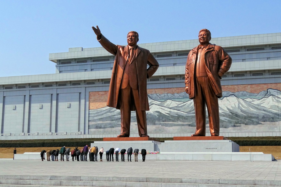 Large statues of two men, both smiling and one is gesturing to the horizon. A long row of people bow before them.