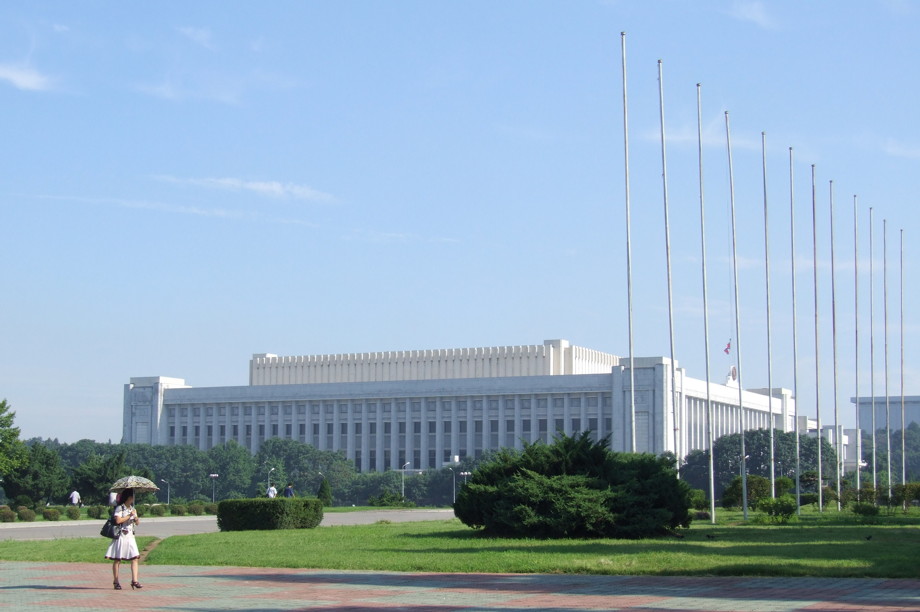 Huge white congressional building in the distance with a line of empty flagpoles to one side and one woman walking by with an umbrella to protect her from the sun
