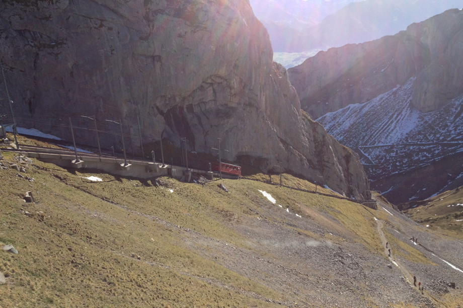 A modest red car going up Mount Pilatus, snow and steep rocks are in the background